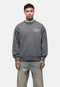 Felpa grigia con colletto rotondo, spalle scese e polsini a coste. Presenta un logo testuale bianco sul petto sinistro. Indossata con jeans di denim chiaro.
