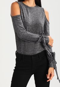 Kvinna i en glittrig silverfärgad topp med kalla axlar och långa ärmar som är knutna vid handlederna, ihop med svarta högmidjade byxor.