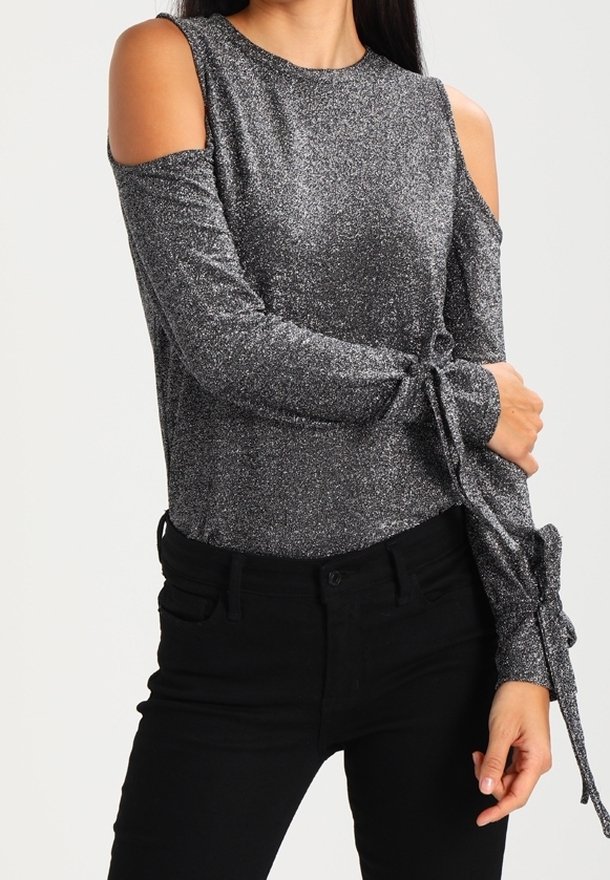 Kvinna i en glittrig silverfärgad topp med kalla axlar och långa ärmar som är knutna vid handlederna, ihop med svarta högmidjade byxor.