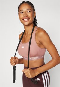 Rosa sportbh med vit "TECHFIT"-band. Svart hopprep hålls i händerna. Enkel design, slät textur och atletisk passform.