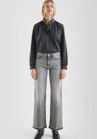 Schwarze Bluse mit langen Ärmeln und gerafftem Halsausschnitt, kombiniert mit hellgrauen weiten Jeans mit fransigem Saum. Schwarze Ankle-Boots runden den Look ab.