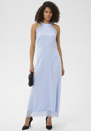Kobieta o kręconych włosach, ubrana w jasnoniebieską, bezrękawnikową sukienkę maxi z koronkowymi zdobieniami, czarne szpilki, srebrną bransoletkę i trzymająca czarną kopertówkę.