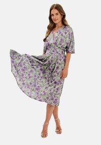 Plynúce zelené šaty s fialovým kvetinovým vzorom, výstrihom do V a krátkymi rukávmi. Majú priliehavý pás a sukňu po členky.