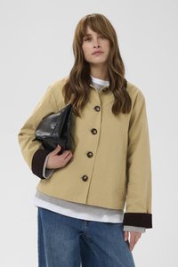 Sieviete ar gariem brūniem matiem, tērpusies bēša krāsas pogātā žaketē, valkā slāņainas pelēkas un baltas virskrekli, zilas džinsas un tur melnu ādas maku.