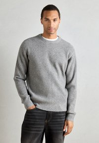 Gray ribbed sweater with a crew neckline and long sleeves, made from a textured knit material. The person wears dark jeans.