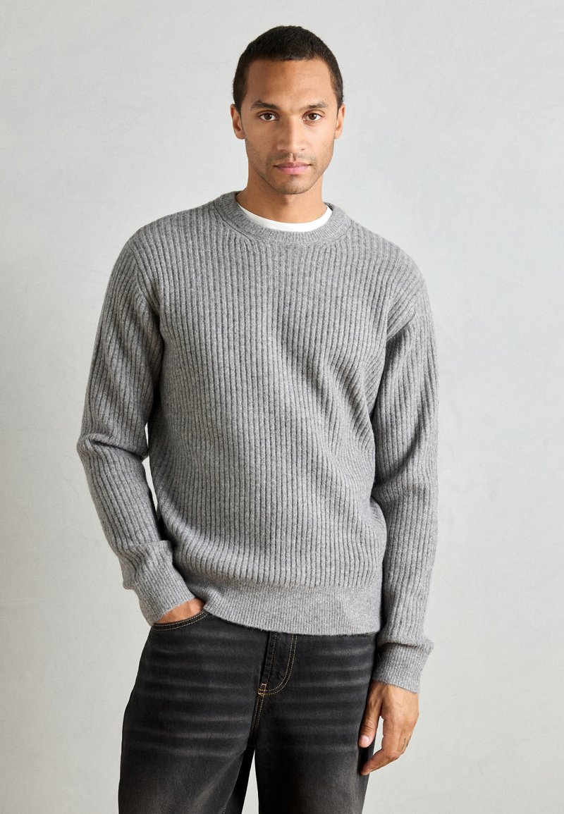 Gray ribbed sweater with a crew neckline and long sleeves, made from a textured knit material. The person wears dark jeans.