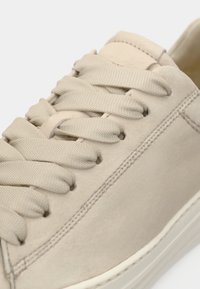 Ανοιχτό μπεζ αθλητικό παπούτσι με λείο επάνω μέρος από σουέντ, στρογγυλεμένη μύτη και επίπεδα κορδόνια. Διαθέτει διακριτικές ραφές και λευκή καουτσούκ σόλα.