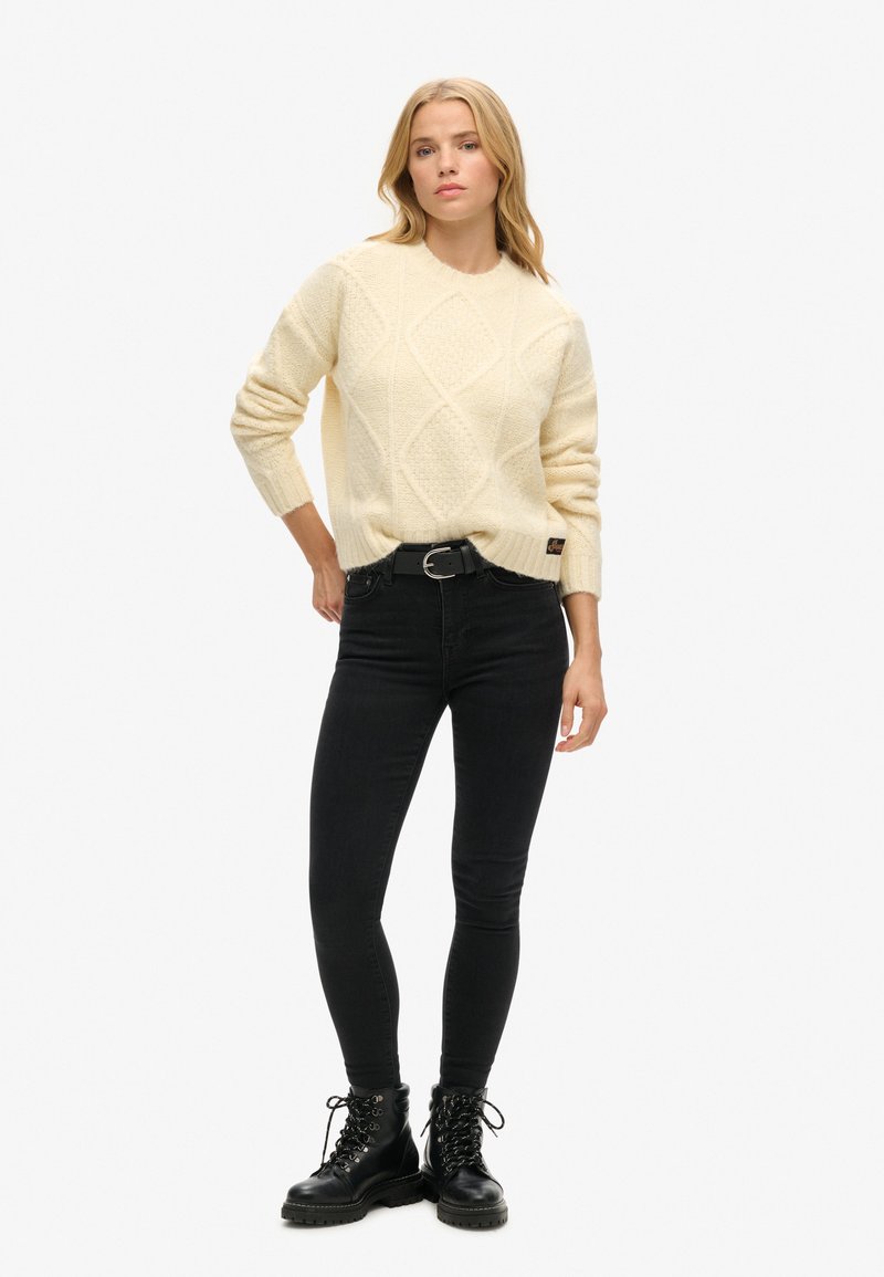 Kremowy sweter z grubego splotu z wzorami w romby, noszony na czarnych obcisłych dżinsach oraz czarnych lace-up butach wojskowych. Prosty czarny pasek jako akcent.