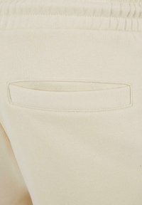 Cream-colored sweatpants feature a smooth texture, elastic waistband, and a small, horizontal back pocket with a stitched border.