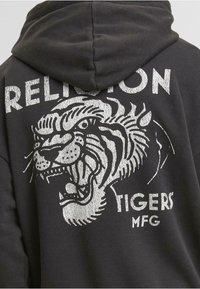 Fekete kapucnis pulóver, ezüst grafikával egy tigris fejével és a "RELIGION TIGERS MFG." felirattal. Puha anyag, laza szabású.