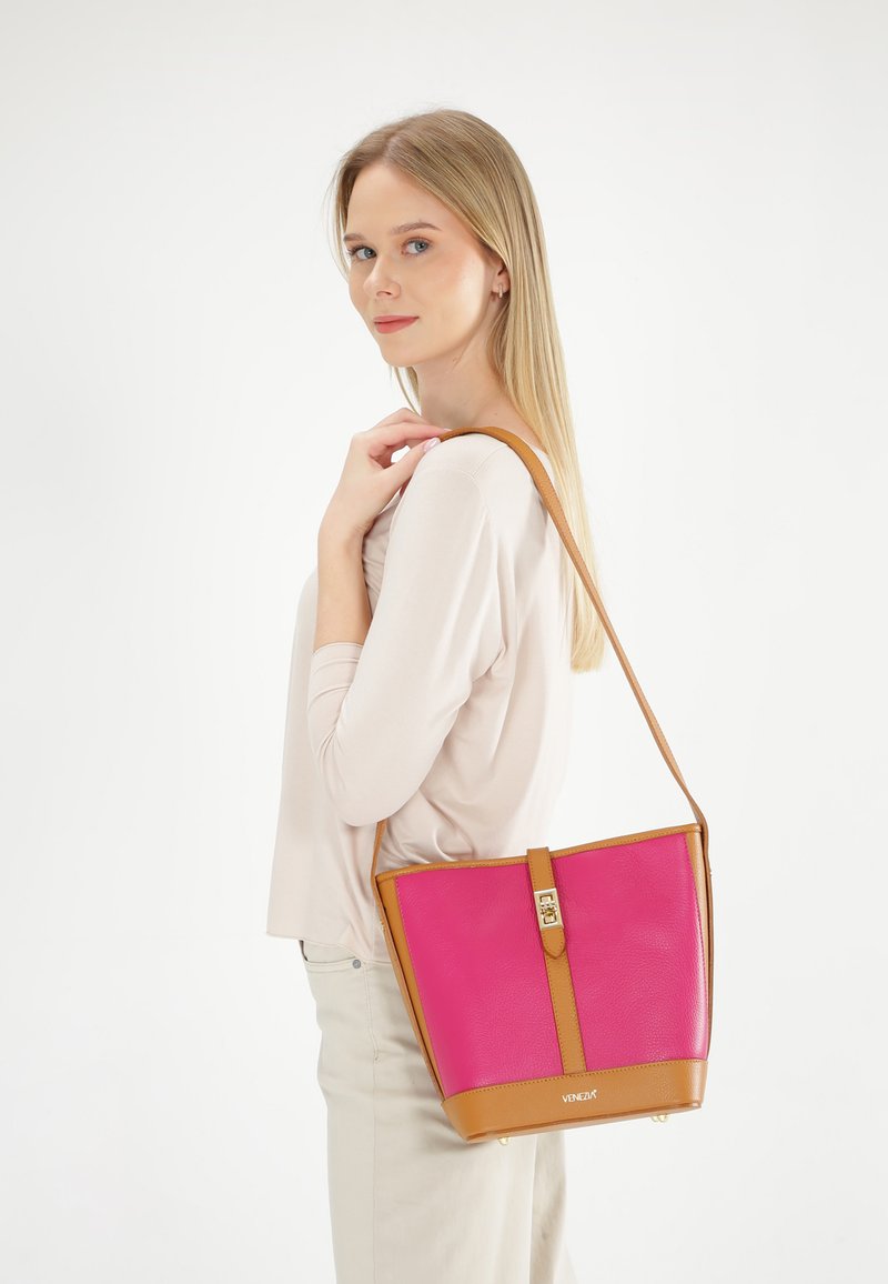 Roze leren handtas met een gladde textuur, bruine accenten, een centrale sluiting en een trapeziumvorm, gedragen over de schouder.