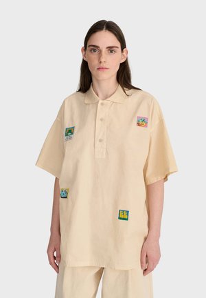 Jeune femme aux longs cheveux foncés portant une chemise beige oversize à boutons avec des patchs colorés sur le devant et un pantalon beige assorti.