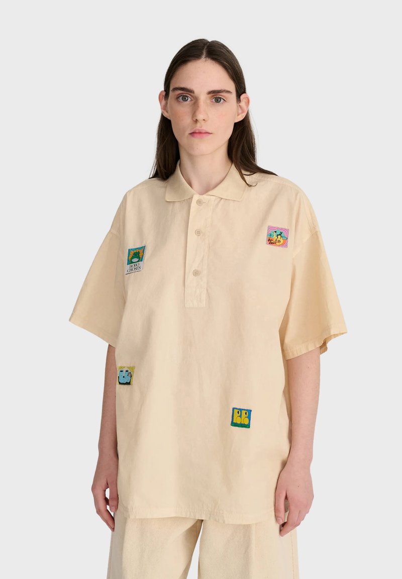 Jeune femme aux longs cheveux foncés portant une chemise beige oversize à boutons avec des patchs colorés sur le devant et un pantalon beige assorti.