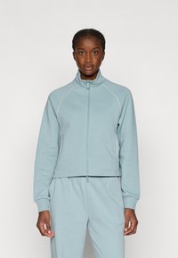 Lyseblå hættejakke med lynlås lavet af blødt stof, med raglanærmer og kontrasterende hvid syning langs sømmene.