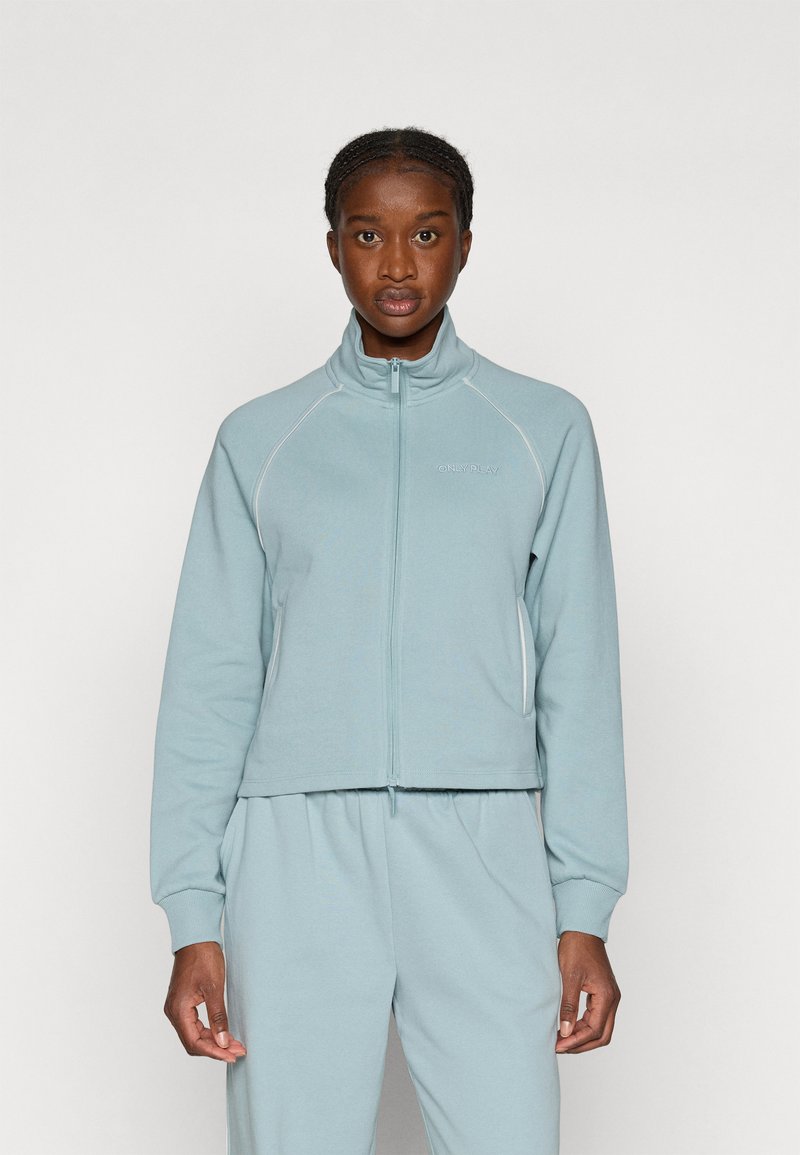 Lyseblå hættejakke med lynlås lavet af blødt stof, med raglanærmer og kontrasterende hvid syning langs sømmene.