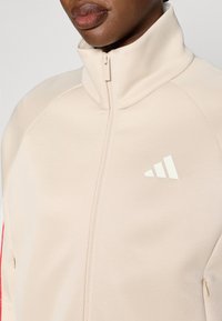 Μπεζ μπουφάν με φερμουάρ και ψηλό γιακά, με λευκό λογότυπο και κόκκι�νες λεπτομέρειες στις πλευρές, κατασκευασμένο από λείο ύφασμα με εφαρμοστή σχεδίαση.