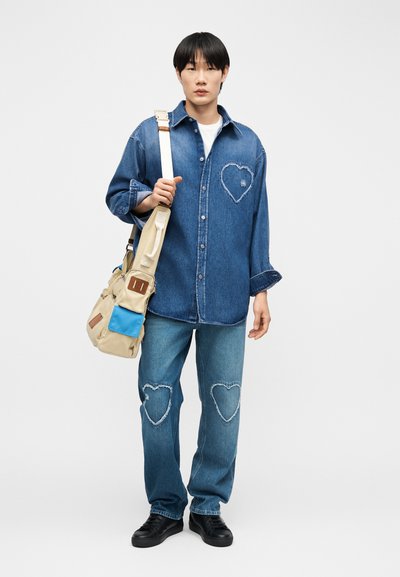 Jeune personne portant une chemise en jean et un jean avec des patchs en forme de cœur, portant un sac à bandoulière beige et bleu, debout devant un fond blanc.