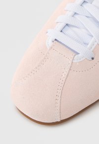 Απαλό ροζ σουέντ αθλητικό παπούτσι με λευκά κορδόνια, στρογγυλεμένο σχήμα μύτης, διακριτικές ραφές και καφέ λαστιχένια σόλα. Ορατή απαλή υφή.