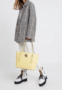 Żółta torba typu tote o gładkiej fakturze, czarnym wykończeniu i metalowym, okrągłym logo. W zestawie z czarno-białą marynarką w wzór pepitki oraz białymi botkami za kostkę.