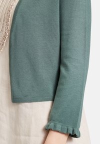 Cardigan corto color teal realizzato in morbido tessuto a maglia con orlo a coste e maniche a sbuffo, indossato sopra un indumento di un leggero color beige.