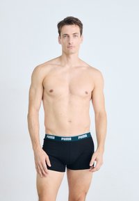 Schwarze Boxershorts mit einem grün-weißen "PUMA"-Logo-Bund. Glattes, eng anliegendes Design aus weichem Stoff. Das Modell posiert frontal.