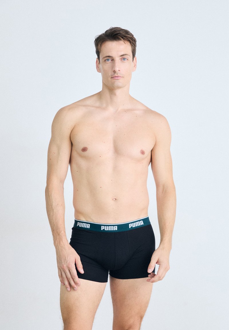 Schwarze Boxershorts mit einem grün-weißen "PUMA"-Logo-Bund. Glattes, eng anliegendes Design aus weichem Stoff. Das Modell posiert frontal.