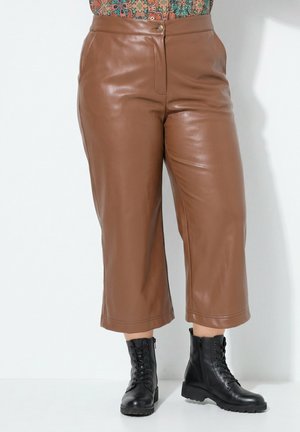 Braune weite, gekürzte Lederhose kombiniert mit schwarzen Schnür-Stiefeletten vor weißem Hintergrund.