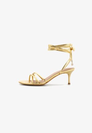 Sandalo dorato con listini, tacco stiletto basso e lacci alla caviglia che terminano con piccoli ciondoli metallici.