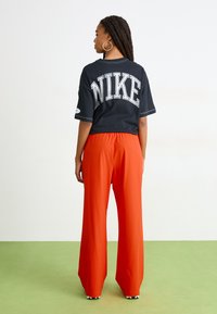 Svart T-skjorte med korte ermer fra Nike med hvitt logo på ryggen, kombinert med løstsittende, høyt liv og lyseoransje bukser. Grønn gulv bakgrunn.