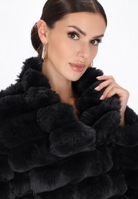 Una donna dai capelli scuri raccolti in uno chignon elegante indossa orecchini a cerchio e un morbido cappotto di pelliccia nera, tenendo il colletto con unghie curate.