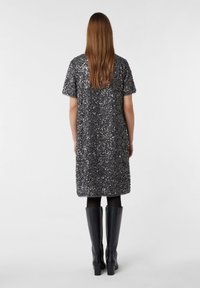 Vrouw met lang bruin haar, gekleed in een knielange, korte mouw donkergrijze sequinedress met zwarte tights en zwarte kniehoog laarzen, met de rug naar de kijker gekeerd.