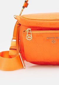 Oranžová crossbody taška s texturovanou tkaninou, zlatým kováním a zipovou přední kapsou. Obsahuje tkaný popruh s nastavitelnou délkou.