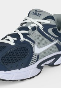 Ναυτικό μπλε και γκρι παπούτσι τρεξίματος Nike με λευκά κορδόνια, διχτυωτά πάνελ και λευκό λογότυπο Swoosh στο πλάι και στη γλώσσα.
