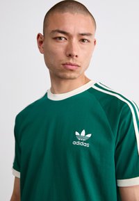 Zöld pamut póló fehér részletekkel és három fehér csíkkal az ujjakon. A mellkason egy kis fehér "adidas" logó található.