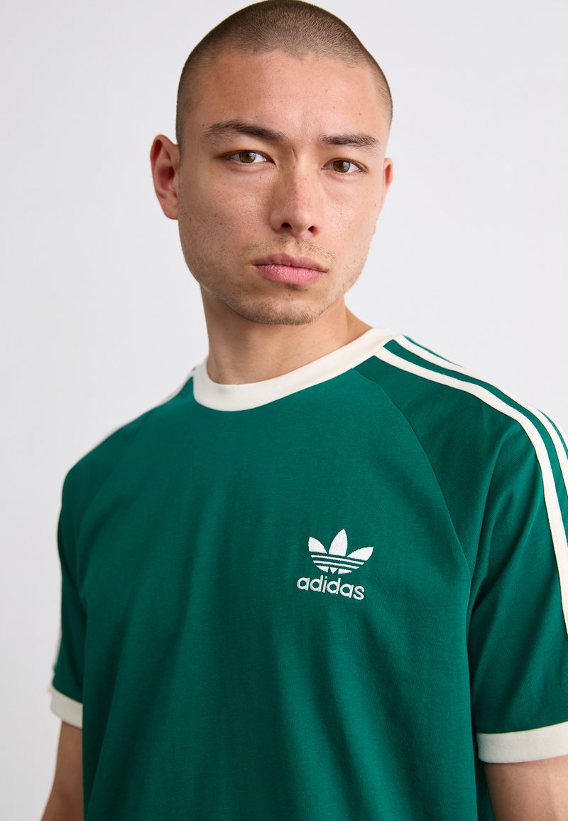 Camiseta de algodón verde con acentos blancos y tres rayas blancas en las mangas. Presenta un pequeño logo blanco de "adidas" en el pecho.
