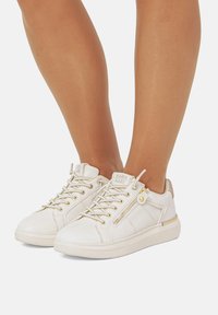 Weiße Sneakers mit strukturiertem Finish, goldenen Akzenten, Schnürsenkeln und einem seitlichen Reißverschluss. Mit einem beigen Absatz und einer Gummisohle.