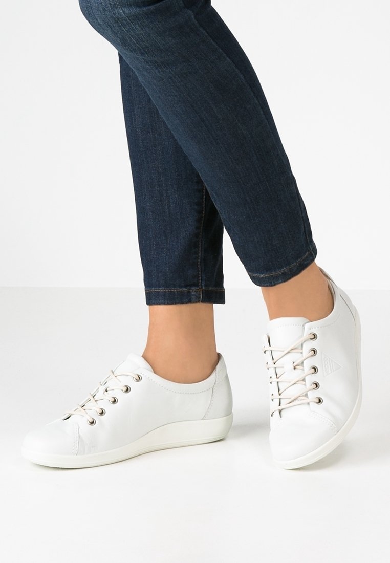 Personne portant un jean bleu foncé et des baskets blanches décontractées à lacets, debout avec un pied légèrement levé.