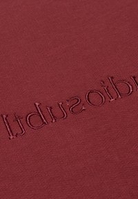 Bordeaux textiel met een textuurstof waarin geborduurde lettering subtiel in het ontwerp is geïntegreerd. Zachte, gladde textuur.
