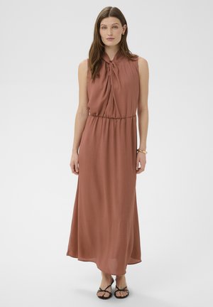 Kobieta ubrana w bezrękawnikową, rdzawego koloru sukienkę maxi z wiązanym dekoltem i czarne sandały na paski, stojąca na jednolitym tle.