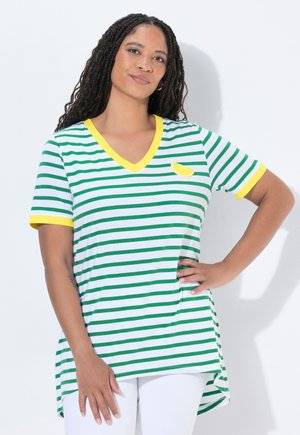 Kobieta z długimi warkoczami, ubrana w zielono-biały pasiasty t-shirt z dekoltem w serek, żółtym wykończeniem i łatką w kształcie cytryny, pozująca z ręką na biodrze.