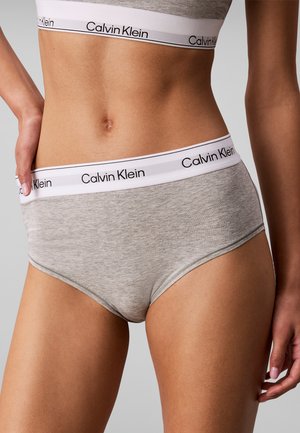 Kobieta ubrana w szary zestaw bielizny Calvin Klein z białymi elastycznymi paskami i czarnym napisem logo, pozująca z ręką na biodrze na jednolitym tle.