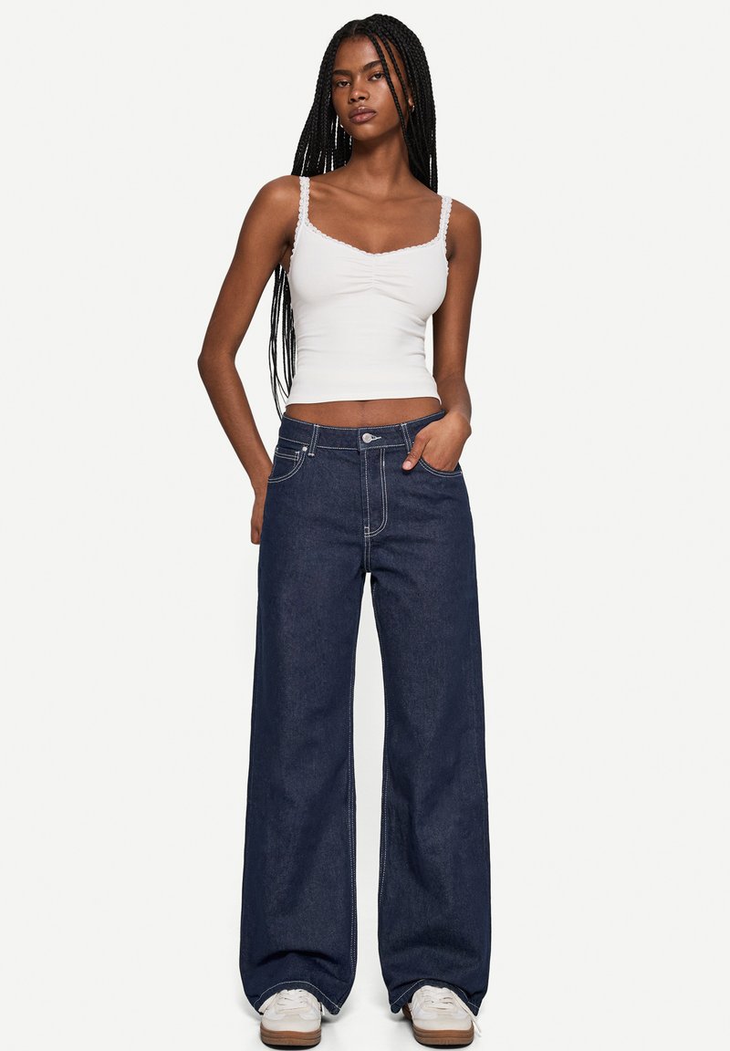 Wide-leg dark denim jeans with contrasting white stitching, featuring a button closure and pockets, paired with a fitted white tank top.