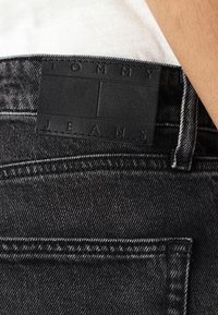 Jeans neri in denim esposti, caratterizzati da un'etichetta in pelle testurizzata sulla vita con la scritta "TOMMY JEANS" in rilievo, insieme a cuciture in contrasto.
