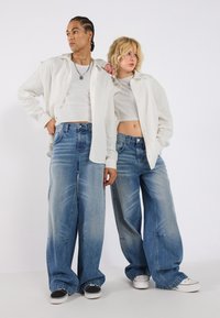 Jean large bleu avec des détails délavés, associés à des tops courts blancs et des chemises à boutons assorties, accompagnés de baskets noires et blanches.