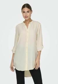 Light cream button-up shirt with a mandarin collar, long sleeves rolled to elbow, and a straight hem. Smooth, lightweight fabric.