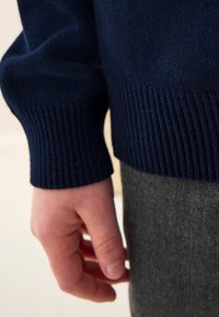 Pull en maille navy avec des poignets côtelés, gros plan sur le détail des poignets, présentant une texture lisse et un design ajusté sur un fond en tissu gris.
