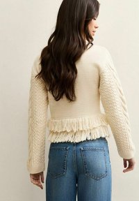 Kobieta o długich ciemnych włosach, ubrana w kremowy sweter z warkoczowym splotem i detalami z frędzlami oraz niebieskie jeansy, stojąca tyłem do kamery.