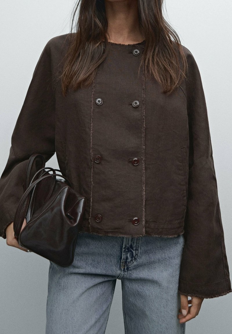 Kvinna i mörkbrun dubbelknäppt jacka och ljusblå jeans, som håller en mörkbrun läderhandväska i vänster hand.