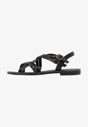 Musta tasapohjainen sandaali, jossa kimaltavat ristikkäiset hihnat ja kaksi säädettävää hopeanväristä solkea sivulla, avoimella varpaalla ja slingback-mallilla.