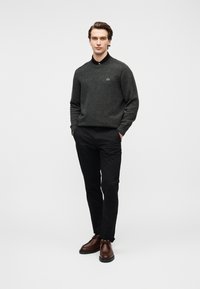 Νεαρός άντρας στέκεται με τα χέρια στις τσέπες, φορώντας σκούρο γκρι πουλόβερ, μαύρο παντελόνι και καφέ δερμάτινα παπούτσια σε λευκό φόντο.